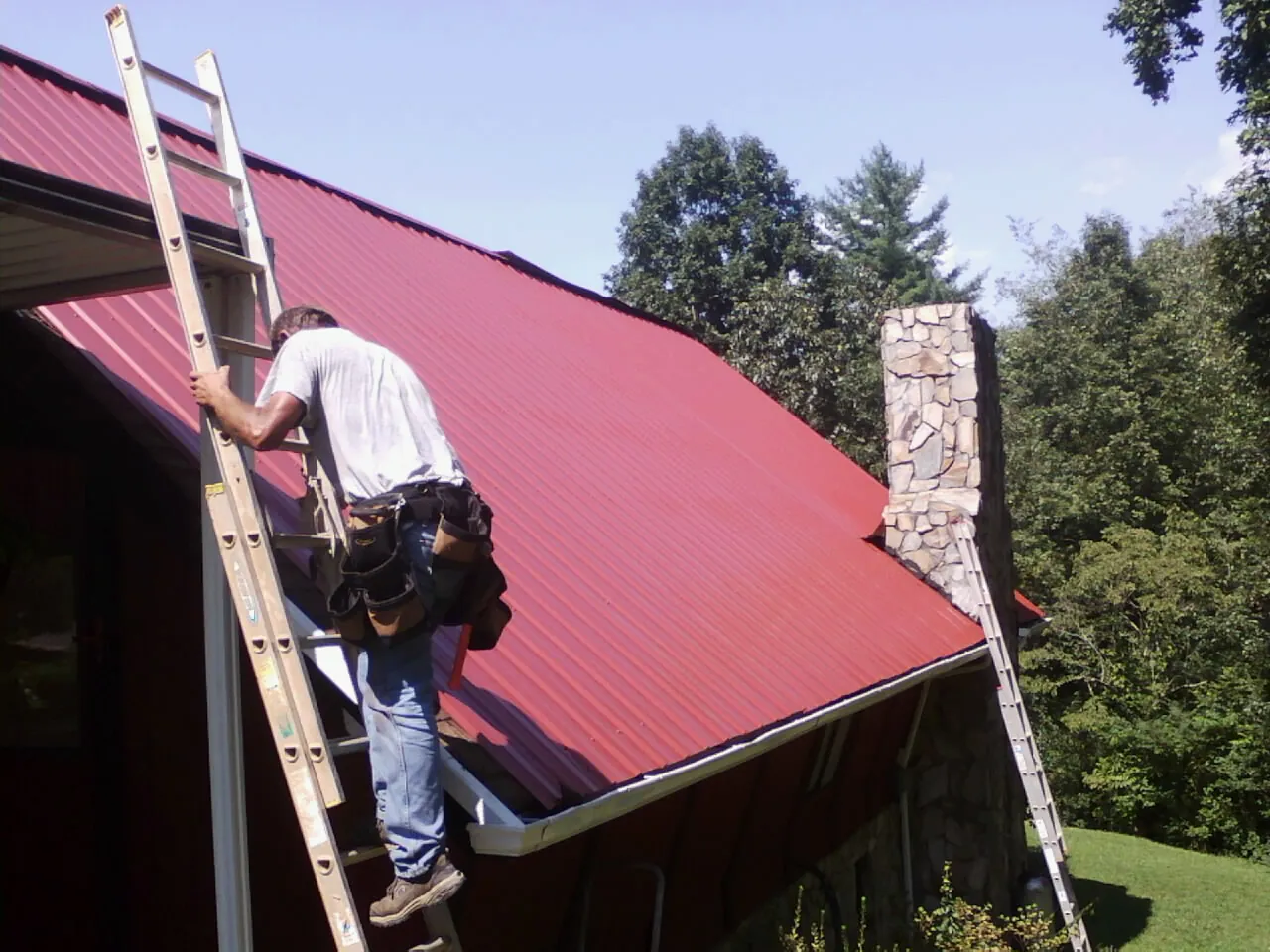 Roofer on a ladder beside a red metal roof and stone chimney — Soffit & Fascia Repair by Roofing Brothers in Clementon